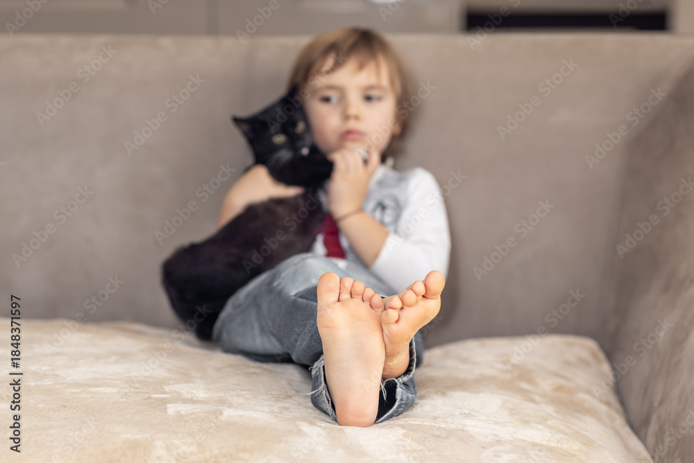 Obraz premium Barefoot child feet on sofa, boy with cat in blurred background