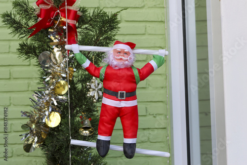 Joyful Christmas decoration featuring red Santa Claus climbing white ladder near green brick wall and pine branch with golden ornament captures festive holiday spirit and merry anticipation