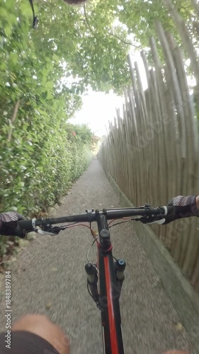 POV. Cyclist rides a mountain bike along a narrow gravel path surrounded by green bushes and wooden fences
