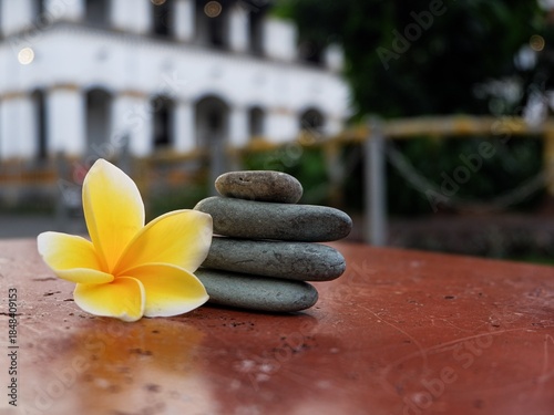 Canvas Print A peaceful Zen composition featuring a stack of four balanced smooth grey river stones next to a yellow plumeria flower on a red surface, with a soft-focus building in the background