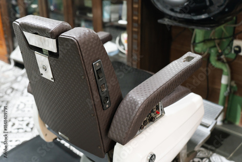 Vintage-style barber chair with brown leather cushion in a classic barbershop interior featuring patterned tile flooring and grooming tools.