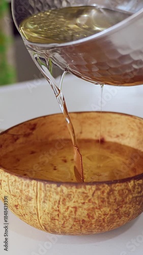 Vertical video. Melted Wax Being Poured from Large Metal Pot into Coconut Bowl on White Table in Soft Natural Light, Handmade Candle Concept