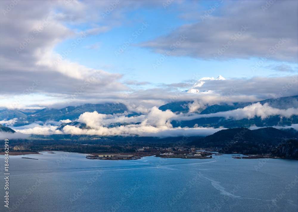 Fototapeta premium Snowy Mountain Peak Rises Over Cloudy Fjord With Calm Water in Squamish Area