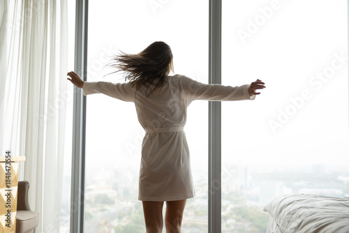 Overjoyed with freedom. Back view carefree millennial woman with slim figure spinning around in bedroom with large picture window admire spectacular city view enjoy fresh daylight in luxury apartment