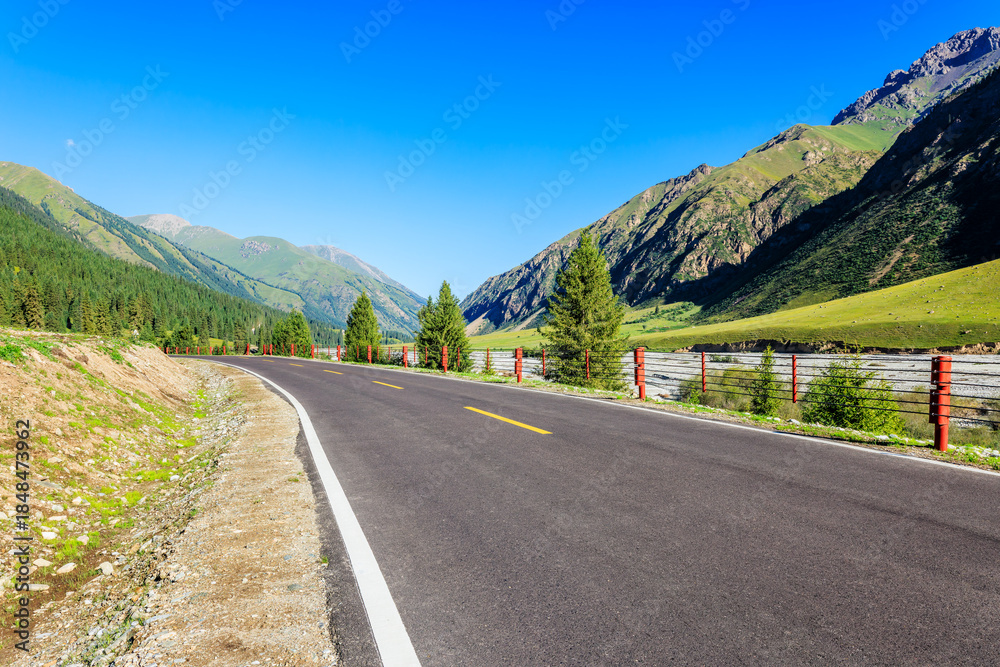 Fototapeta premium Empty asphalt road winding through green mountains and valley under clear blue sky in summer