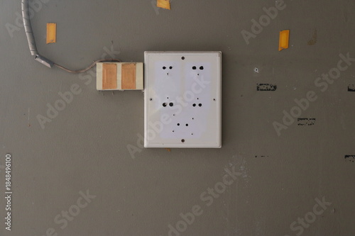 An abandoned load center of a closed down shop. It is empty showing only mounting holes. To the left, an electrical outlet masked with adhesive tapes is visible with cable running into it.