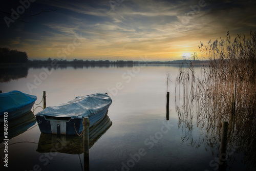 A tranquil lakeside scene at sunset with a small moored boat resting on calm water, surrounded by reeds and wooden posts