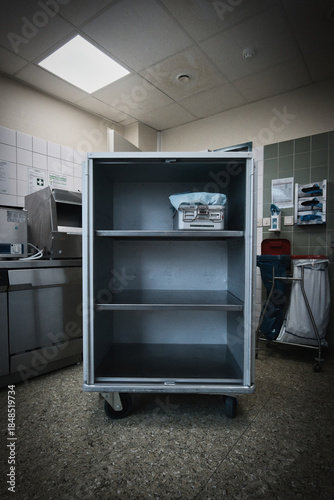 An empty stainless steel hospital storage cart with shelves, standing in a clinical environment.