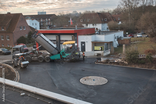 An elevated view of an active road construction site in a residential urban area, featuring asphalt paving machinery, a dump truck, and construction workers wearing safety gear.