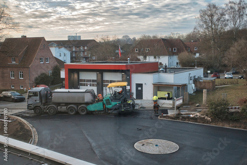 An elevated view of an active road construction site in a residential urban area, featuring asphalt paving machinery, a dump truck, and construction workers wearing safety gear