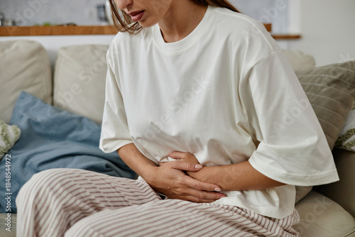 Young adult Caucasian woman sitting on sofa holding stomach with both hands appearing to experience abdominal pain or discomfort, upper body and face partially visible