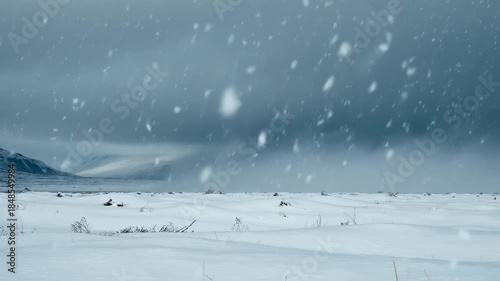 A snowy landscape features falling flakes, snowy field, hills and a dramatic overcast sky