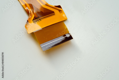 Macro shot of an orange flexible printed circuit (FPC) cable on a white background. Electronic component showing gold contact pins and internal traces for data transmission.