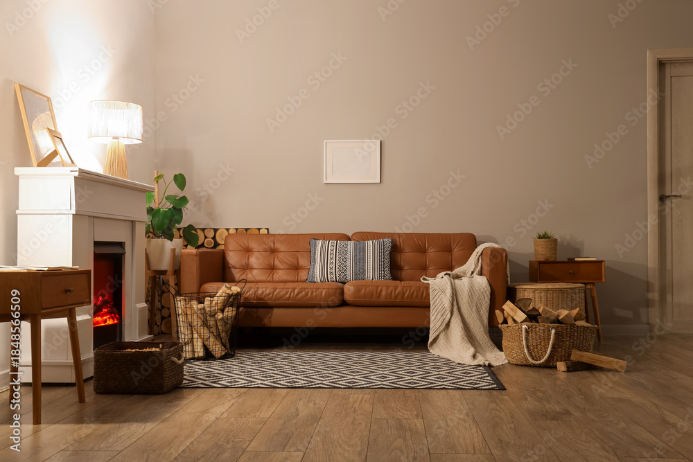 Fototapeta premium Interior of living room with sofa, glowing lamps, bedside tables and firewood near fireplace in evening