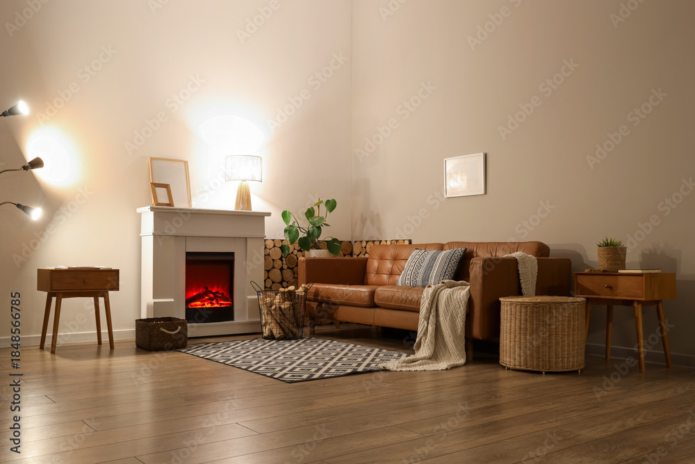 Obraz premium Interior of living room with sofa, glowing lamps, bedside tables and firewood near fireplace in evening