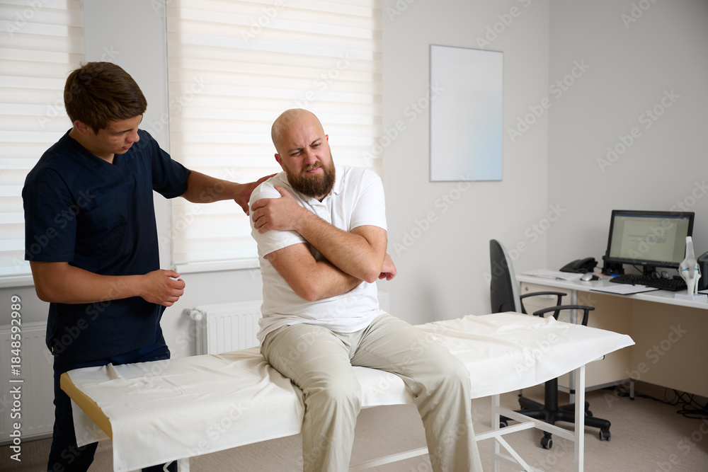 © Василь Івасюк - Physiotherapist examining patient shoulder joint during rehabilitation therapy session in modern medical clinic. Man receiving orthopedic treatment and physical therapy for injury recovery.
