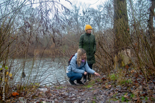 Fotografi Two women exploring a damp, wintry lakeside, one crouching to examine the leaf-strewn ground