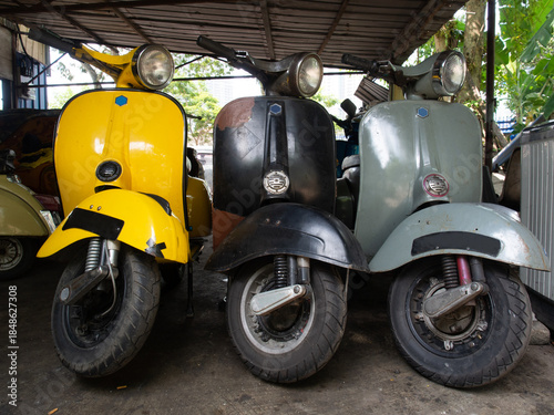 Three classic retro scooter in yellow, black and grey parked under a metal sheet awning shade.