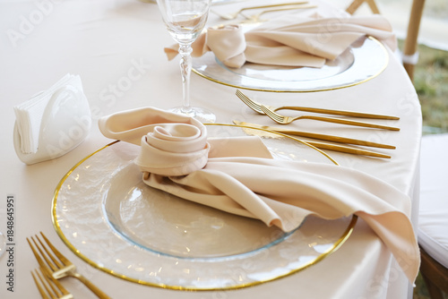 Elegant table setting with gold cutlery, glassware, and folded napkins on white tablecloth, featuring clear plates with gold rims and decorative elements arranged neatly