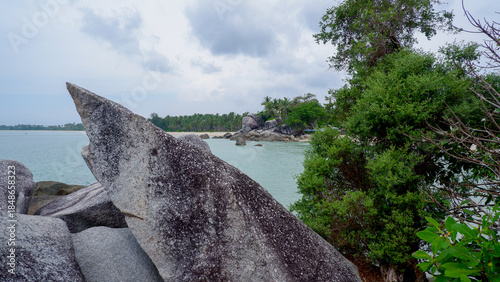 Distinctive sharp granite rock formation on a tropical rocky coastline with calm sea, green vegetation, and cloudy sky.