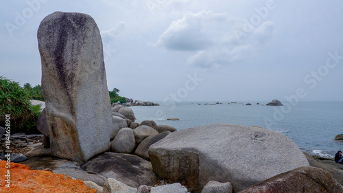 Tall standing granite boulder on a tropical rocky beach with red sand foreground, calm sea, and cloudy sky.