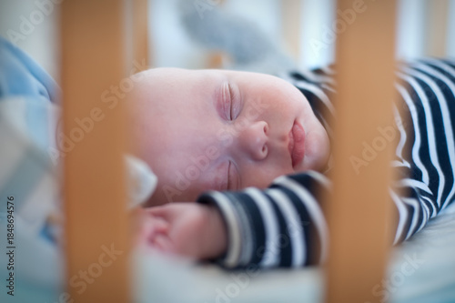 An infant is peacefully asleep in a crib. The baby wears a striped shirt and is resting on a white sheet, behind the wooden bars of the crib. The afternoon light is soft.