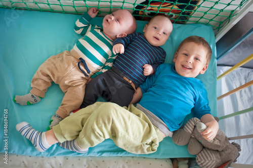 Three young brothers are laying in their baby bed. Two are infants and one is a young boy. They are wearing casual clothing and socks. One boy smiles while holding a stuffed animal.