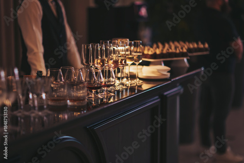 People socialize at a bar with different types of drinks arranged neatly on the counter near a selection of snacks. The event takes place during the evening in a stylish venue