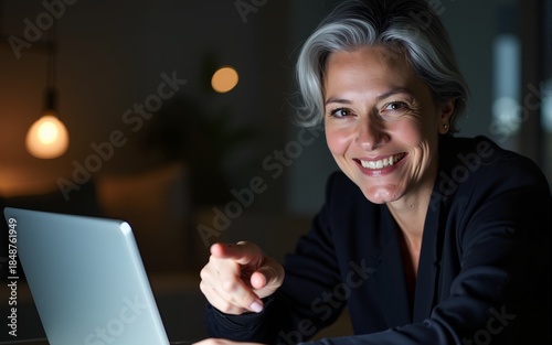 Middle age woman with grey hair working using computer laptop late at night smiling and looking at the camera pointing with two hands and fingers to the side. High quality