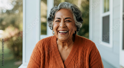Smiling elderly woman with gray hair wearing an orange sweater, sitting outdoors on a porch, radiating joy and warmth in a serene environment filled with natural light