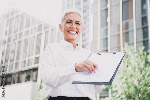 Tableau sur toile Confident senior woman in white striped shirt holding clipboard smiles outside m