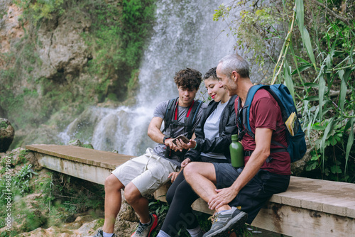 Family hikers resting by waterfall checking smartphone