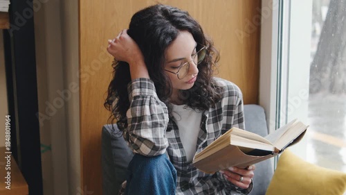 Woman sitting by a window during daytime reading a book with a calm expression in a cozy indoor space