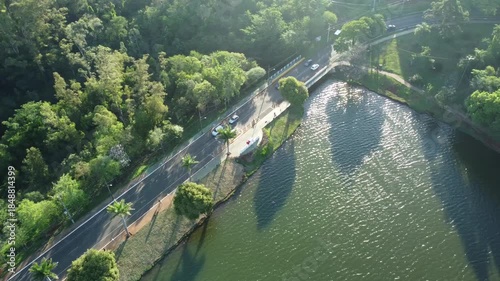 Aerial view of a road and bridge beside an urban lake, surrounded by green vegetation. Sunlight reflects on the water, showing the connection between city infrastructure and nature.