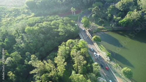 Aerial view of a road and bridge beside an urban lake, surrounded by green vegetation. Sunlight reflects on the water, showing the connection between city infrastructure and nature.