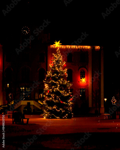 A stunningly large Christmas tree with fairy lights stands in a dark night in a public square with park benches in front of a building with a clock and a staircase. Vertical photograph.
