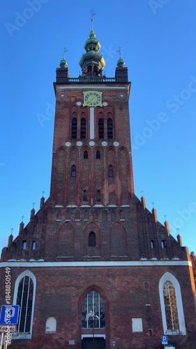 St Catherine's Church is the oldest church in Gdansk, Poland. A tall brick church tower rises against a clear blue sky. The clock shows the time while sunlight falls on the structure.