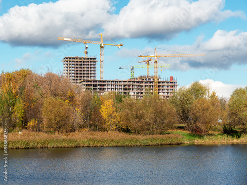Residential building construction site with high tower cranes under white clouds and blue sky view from autumn riverbank