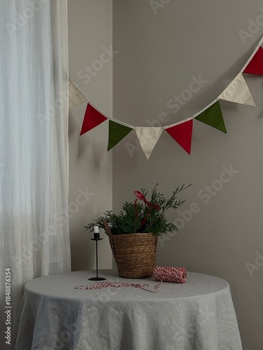 Cozy Table With Bunting, Basket Plant. Soft Natural Light Falling Through Sheer Curtain, Neutral Linen Tablecloth Draped Over Round Table, Small Vintage Lamp Casting Warm Glow,