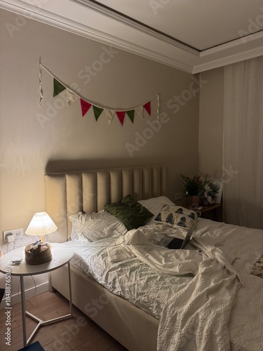 Cozy Bedroom With Unmade Bed, Bedside Lamp Glow. Neutral Linen And Textured Blanket Draped Across Mattress, Colorful Bunting Above Radiator, Small