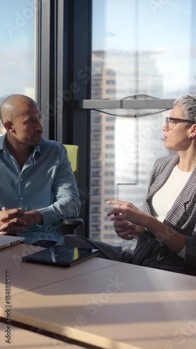 Vertical. A male and female coworker analyze work on a laptop beside a large window with skyline views, highlighting problem solving, coordination, and an efficient workflow in a contemporary office.
