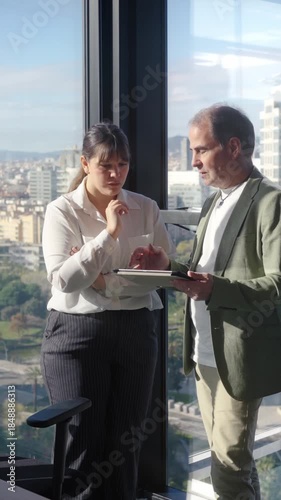 Vertical. Colleagues review a tablet together by the window, with the manager offering mentoring and clear guidance during a one to one discussion inside a bright contemporary office.