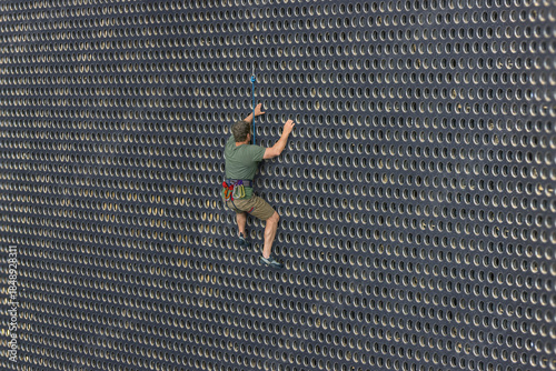 A climber with a safety rope climbs a circular perforated metal wall