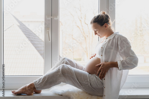 Side view of beautiful pregnant woman holds hands on belly by the window at home. Pregnancy, maternity, preparation and expectation concept. Beautiful tender mood photo of pregnancy.