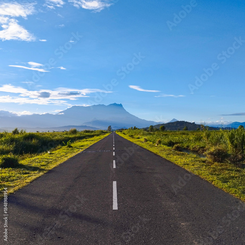 A scenic perspective of the Tempasuk Plains, where a long highway points directly toward the granite massif of Mount Kinabalu, a UNESCO World Heritage site.