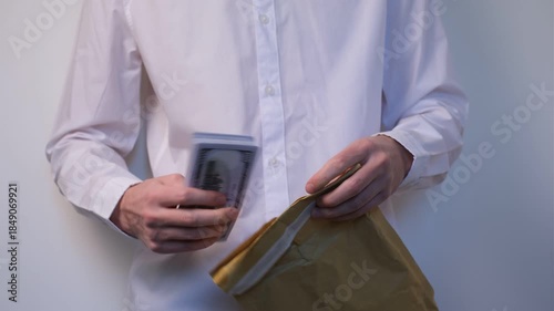 Young man in white shirt counts stack of one hundred dollar bills and puts them into craft paper bag on white background