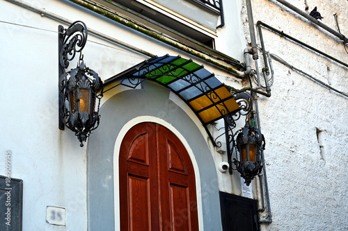 arched brown stained entrance door detail with decorative metal and color glass canopy above. antique wall sconces on the side. balcony above. electric cable conduits. white stucco elevation