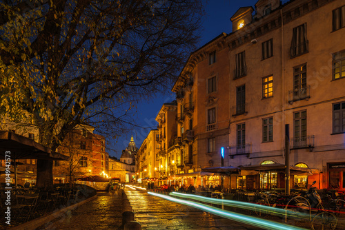 Turin, Italy. Night view of Turin Duomo from Largo IV Marzo, with glowing street lights, outdoor cafes, people and car light trails, capturing historic architecture and urban nightlife.
