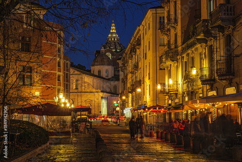 Turin, Italy. Night view of the Cathedral from Largo IV Marzo, with warm street lights, outdoor restaurant tables, historic buildings and people creating a lively evening city atmosphere.
