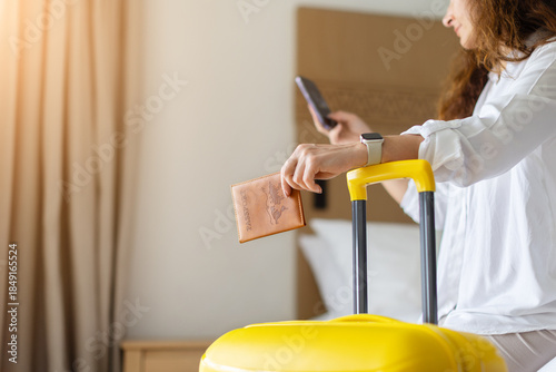 Quadro em tela Adult woman holding passport and smartphone beside yellow suitcase in home bedroom